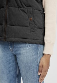 Svart vadderad väst med dragkedja och en ficka med tryckknapp, bärs över en ljusfärgad tröja; kombinerad med ljusblå jeans.