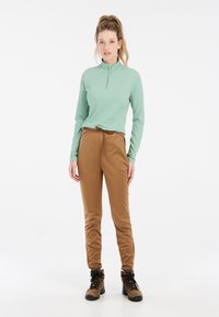 Maglia a maniche lunghe verde chiaro con zip abbinata a pantaloni beige con vita elastica. Il modello indossa scarponi da trekking marroni, mostrando un look elegante e pronto per l'outdoor.