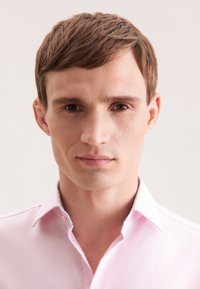 Giovane uomo con capelli corti castani e occhi marroni, che indossa una camicia a maniche lunghe di colore rosa chiaro, con uno sguardo neutro rivolto dritto davanti a sé.