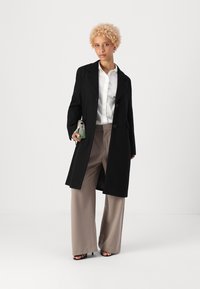 Cappotto di lana nero con una vestibilità sartoriale, abbinato a una camicia bianca con bottoni e pantaloni beige a gamba larga. Borsa piccola in tessuto e scarpe con punta aperta.