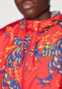 Windbreaker rojo con patrones abstractos en azul y amarillo, con cremallera en el frente, cordones en el cuello y un logo de FCB en el pecho.