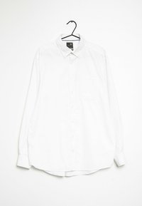 Biała, długa koszula z długim rękawem zawieszona na czarnym wieszaku. Posiada wyłogi, kieszonkę z przodu oraz miękką, gładką tkaninę.