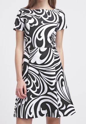 Femme portant une robe à manches courtes noire et blanche avec des motifs tourbillonnants abstraits audacieux, debout devant un fond uni.