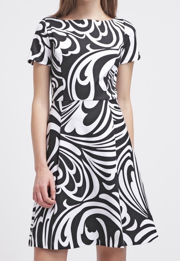 Femme portant une robe à manches courtes noire et blanche avec des motifs tourbillonnants abstraits audacieux, debout devant un fond uni.
