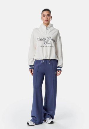 Naine, kes kannab valget veerandlukuga dressipluusi tekstiga "Cardio Bunny Club", tumesiniseid laiade säärega pükse ja valgeid tenniseid, seisab valge tausta ees.