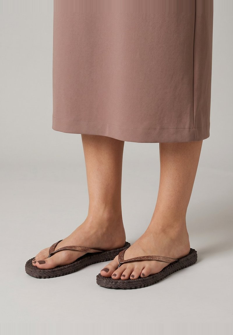 Pieds portant des sandales marron avec semelles texturées et vernis à ongles marron, visibles sous une jupe marron longueur genou sur un fond clair.