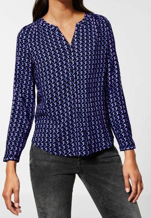 Mujer con blusa de manga larga con un estampado de formas geométricas en azul, rosa y negro, combinada con jeans negros deslavados.