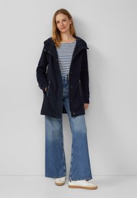 Marineblauer Kapuzenmantel mit Reißverschlusstaschen, über einem gestreiften Oberteil und weit geschnittenen blauen Jeans, kombiniert mit weißen Sneakers und hellbraunen Sohlen.