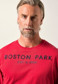 Rotes T-Shirt aus Baumwolle mit marineblauem, gesticktem Text "BOSTON PARK EST 1996." Rundhalsausschnitt und kurze Ärmel. Glatte Textur.