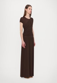 Brun maxi-kjole med en tettsittende bodice som har draperte detaljer og korte ermer, som flyter ned til en løs skjørt laget av glatt stoff.