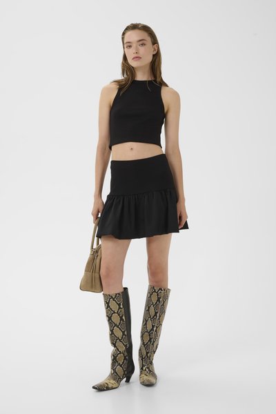 Femme portant un crop top noir sans manches et une mini-jupe à volants, tenant un sac beige, avec des bottes hautes à motif peau de serpent, sur fond blanc.