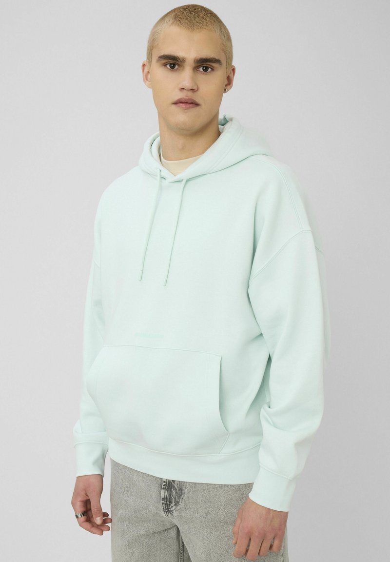 Jeune homme aux cheveux blonds courts portant un sweat à capuche bleu clair et un jean gris, debout devant un fond gris uni.