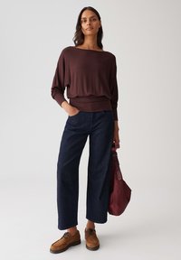 Braunes, locker sitzendes Oberteil mit weitem Ausschnitt, dunkelblaue Schlagjeans und braune Schuhe. Das Model hält eine burgunderfarbene Handtasche. Einfaches, lässiges Outfit.