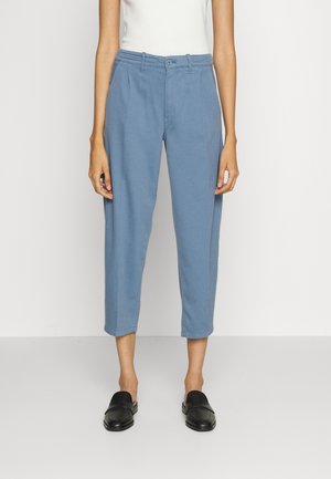 Vrouw draagt blauwe cropped broek met voorplooien, witte mouwloze top en zwarte instap loafers tegen een effen achtergrond.