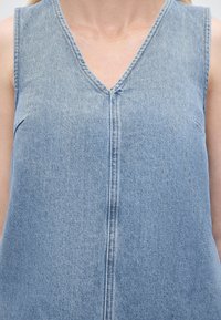 Αμάνικο ανοιχτό μπλε φόρεμα από τζιν με λαιμόκοψη σε σχήμα V και κεντρικό ραφή, φορεμένο από άτομο με ανοιχτόχρωμο δέρμα και ξανθά μαλλιά.