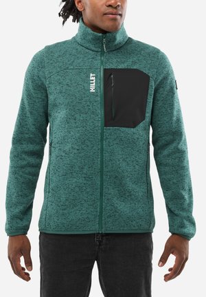 Veste en polaire verte avec fermeture éclair, col montant et poche poitrine zippée noire. Caractéristiques de tissu texturé et de accents de fermeture éclair noire.