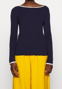 Osoba oblečená v námořnickém modrém svetru s dlouhým rukávem a bílým lemem na výstřihu a manžetách, doplněná o jasně žluté kalhoty s vysokým pasem.