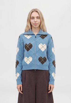 Une femme porte un cardigan bleu zippé orné de motifs de cœurs crème et bruns, assorti à un pantalon à carreaux bruns, sur un fond uni.