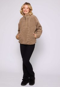 Uśmiechnięta kobieta stojąca z rękami w kieszeniach, ubrana w brązowy polar, czarne spodnie i czarne buty na jednolitym tle.