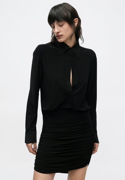 Juoda ilgomis rankovėmis marškiniai su sagomis prie apykaklės ir centrinėmis skeltukėmis, derinami su aptempta raukšlėta juoda mini sijonu. Lygios audinio tekstūros.