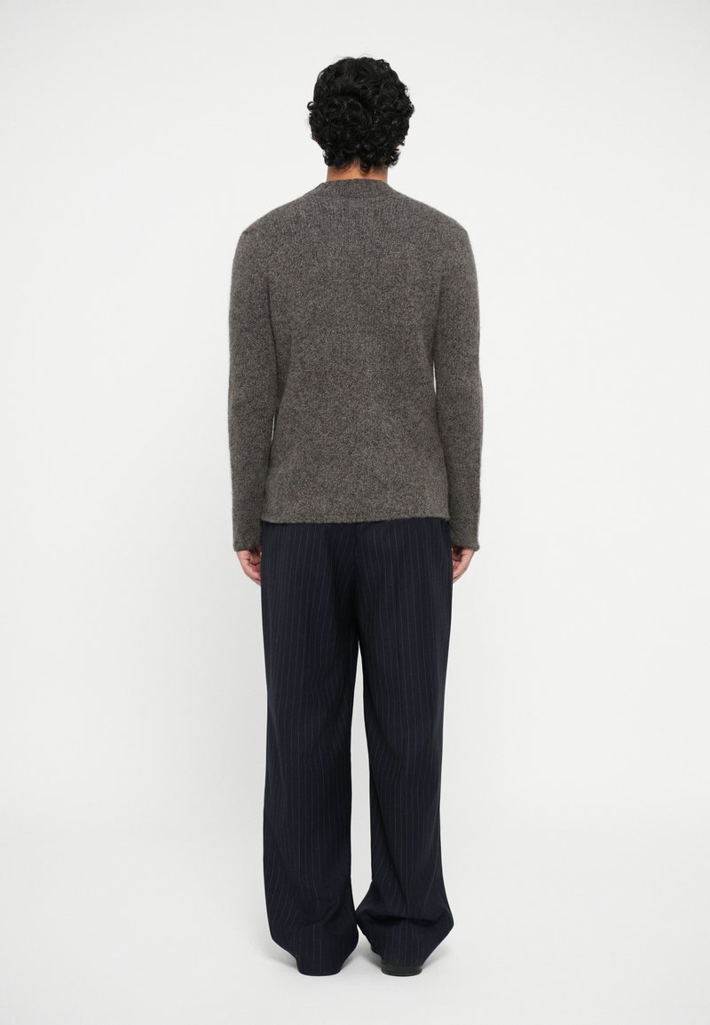 Maglione marrone lavorato a maglia con maniche lunghe, texture grigiastra, abbinato a pantaloni larghi a righe scure. Il tessuto appare morbido e caldo.