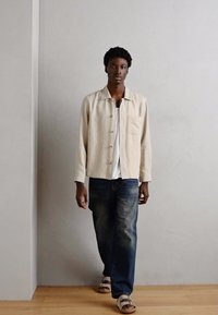 Beige vászon ing, mellzsebbel, sötétkék, laza szabású farmernadrággal és barna szandállal párosítva, minimalista háttér előtt.