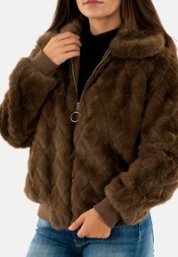 Giacca in pelliccia sintetica marrone con texture trapuntata, chiusura con zip e polsini a coste. Presenta un colletto morbido e una vestibilità aderente e corta.