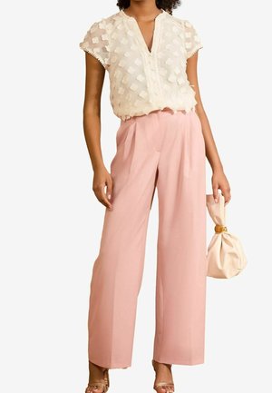 Kobieta ubrana w białą, teksturowaną bluzkę z krótkim rękawem, włożoną do wysokich, szerokich różowych spodni, trzyma kremową torebkę z marszczeniem.