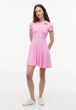 Frau steht und trägt ein rosa Polo-Kleid mit kurzen Ärmeln, ausgestelltem Rock und weißen Sneakers vor einem schlichten weißen Hintergrund.