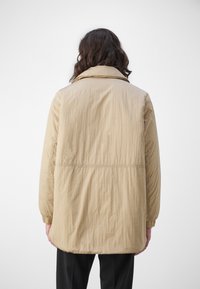 Lichte beige jas met een gestructureerde afwerking, met een lange silhouette, brede kraag en elastische manchetten. De achterkant toont een eenvoudig ontwerp.