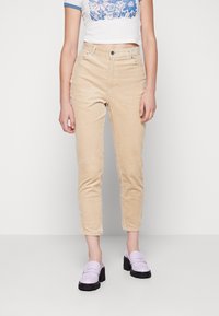 Pantalons en velours côtelé beige avec coupe droite, dotés de poches avant et d'un zip. Portés avec un crop top graphique blanc et des mocassins violet clair.