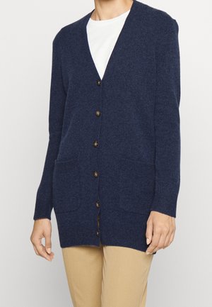 Femme portant un cardigan bleu marine boutonné avec des poches sur le devant, par-dessus une chemise blanche et un pantalon beige, sur un fond uni.