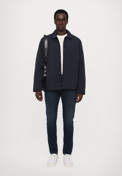 Marineblauer gesteppter Mantel mit Druckknöpfen, getragen über einem weißen strukturierten Pullover, kombiniert mit dunklen Jeans und weißen Sneakern. Schwarze Umhängetasche.
