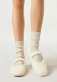 Weiße, flauschige Hausschuhe mit einer abgerundeten Zehenpartie und einem Riemen über dem Spann. Getragen mit hellbeigen, gerippten Socken, die dezente Glitzerakzente aufweisen.