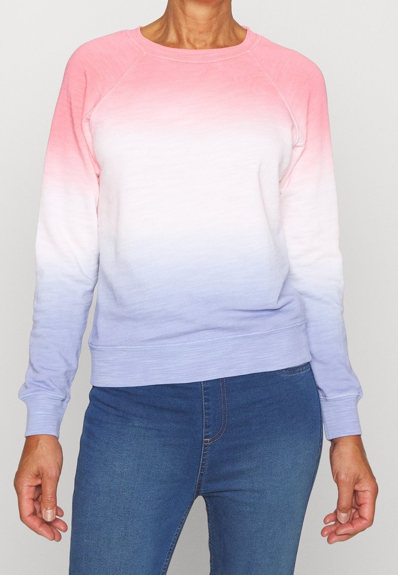 Donna con una felpa a maniche lunghe e colletto rotondo, caratterizzata da una sfumatura di rosa, bianco e viola, abbinata a jeans in denim blu.