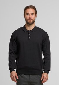 Mann mit Bart, der einen schwarzen Langarm-Polo-Pullover mit drei Knöpfen trägt, stehend vor einem einfarbigen hellgrauen Hintergrund.