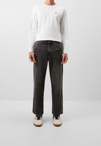 Camisa blanca de manga larga con un pequeño logo rojo, combinada con pantalones vaqueros de pierna ancha gris oscuro y zapatillas beige con acentos azules.