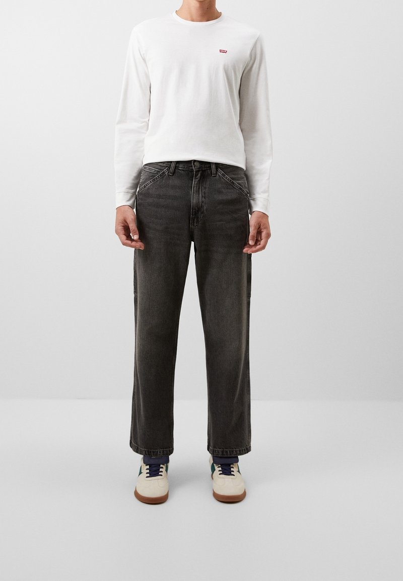 Camisa blanca de manga larga con un pequeño logo rojo, combinada con pantalones vaqueros de pierna ancha gris oscuro y zapatillas beige con acentos azules.