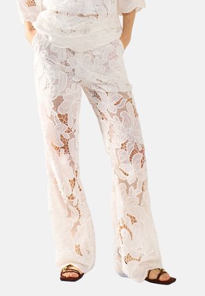 Vrouw draagt witte brede broek van bloemrijke kant met zijzakken en bruine sandalen met open teen, staand met handen in de zakken.