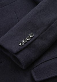 Manga de chaqueta de lana azul marino con cuatro botones negros y un sutil detalle de costura. Tejido suave y texturizado con una forma ajustada.