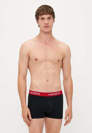 Schwarze Boxer Shorts mit rotem Bund, versehen mit dem "LACOSTE"-Logo, aus glattem Stoff gefertigt und für Komfort und Passform entworfen.