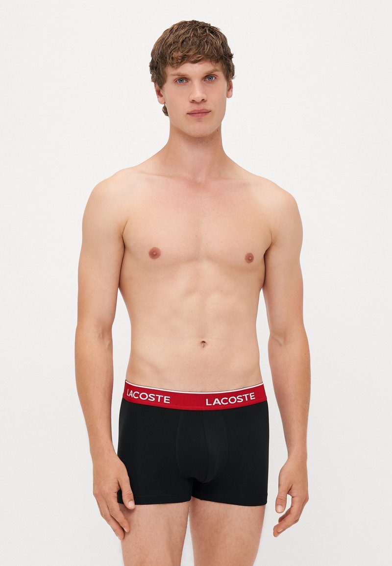 Boxer-briefs noirs avec une taille rouge portant le logo "LACOSTE", fabriqués en tissu doux et conçus pour le confort et l'ajustement.