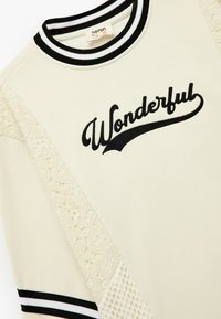 Beige sweatshirt met zwart-wit gestreepte ribboord, geborduurde "Wonderful", en kant- en gaasaccenten op de zijpanelen.