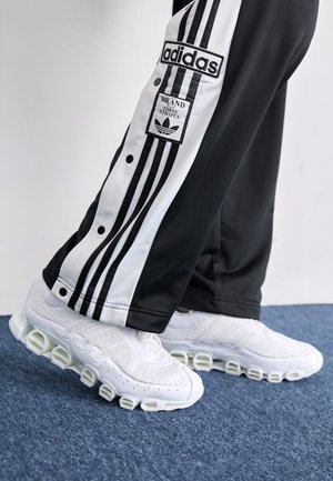 Persoon in zwarte Adidas-broek met witte strepen en witte Adidas-sneakers die op een blauwe vloer van tapijt staat.