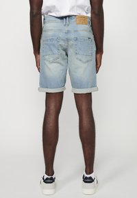 Des shorts en denim bleu clair présentent un ourlet roulé, deux poches arrière et une étiquette en cuir beige à la taille. Portés avec des baskets blanches.