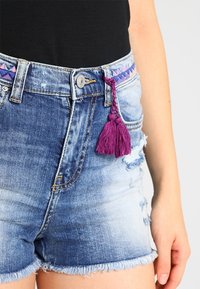 Jeansowe szorty w jasnoniebieskim kolorze, z postrzępionymi brzegami, przecieranymi detalami oraz fioletową ozdobą w postaci frędzli na pasku.