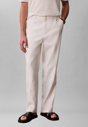 Mężczyzna w jasnobeżowych spodniach, beżowej koszuli z krótkim rękawem, czarnych skórzanych sandałach, stoi z rękami w kieszeniach na szarym tle.