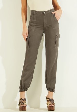 Mujer con pantalones cargo de talle alto en verde oliva con puños elásticos y sandalias de plataforma con estampado animal, de pie sobre un fondo claro y liso.