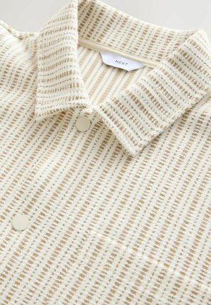 Krems krāsas krekls ar brūniem vertikāliem svītrām, ar gludu audumu un divām saskaņotām pogām pie apkakles. Redzama zīmola birka.