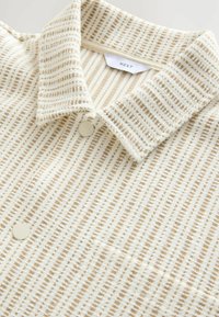 Chemise rayée crème et beige avec un col classique, dotée d'un tissu texturé, d'une fermeture à deux boutons et de motifs subtils tout au long.